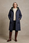 A long quilted hooded coat in dark navy blue with gold-tone snap buttons and black trim, hanging against a light gray background.