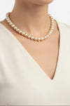 A strand of irregular baroque pearls in a warm champagne-gold color, arranged in a circle on a light cream background. The pearls have a glossy, lustrous finish and are secured by a small dark metal clasp.