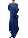 A royal blue two-piece dress displayed on a mannequin, featuring an asymmetrical draped off-the-shoulder top and a pleated high-low skirt. The mannequin is styled with a matching blue velvet crossbody bag with gold hardware, a gold chain necklace, and black boots, set against a home interior background with a door and curtain.