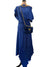 A royal blue two-piece dress displayed on a mannequin, featuring an asymmetrical draped off-the-shoulder top and a pleated high-low skirt. The mannequin is styled with a matching blue velvet crossbody bag with gold hardware, a gold chain necklace, and black boots, set against a home interior background with a door and curtain.