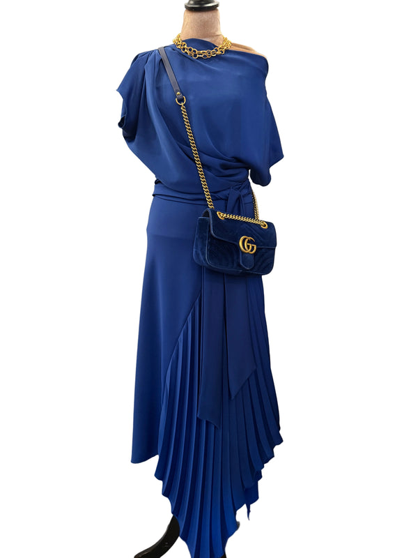 A royal blue two-piece dress displayed on a mannequin, featuring an asymmetrical draped off-the-shoulder top and a pleated high-low skirt. The mannequin is styled with a matching blue velvet crossbody bag with gold hardware, a gold chain necklace, and black boots, set against a home interior background with a door and curtain.