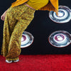 Person wearing yellow floral pants and a mustard jacket on a red carpet with a black background featuring silver circular designs.